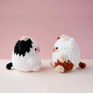 May include: Two crocheted stuffed toys shaped like cows. One cow is white with black spots and a brown nose. The other cow is white with brown spots and a pink nose. Both cows are looking to the right.