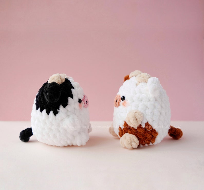 May include: Two crocheted stuffed toys shaped like cows. One cow is white with black spots and a brown nose. The other cow is white with brown spots and a pink nose. Both cows are looking to the right.