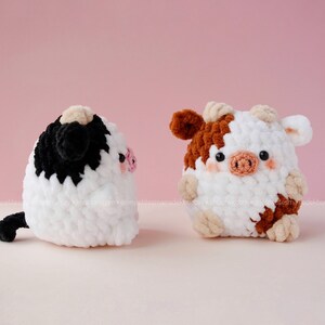 May include: Two crocheted cow plushies, one black and white and one brown and white, with pink noses and black eyes. The plushies are sitting on a white surface.