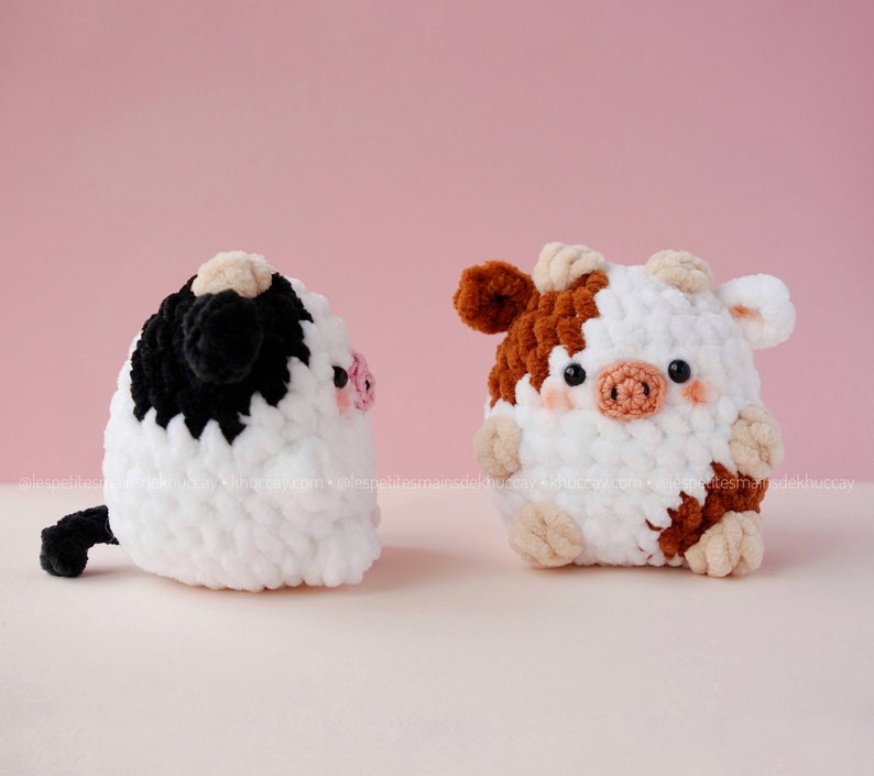 May include: Two crocheted cow plushies, one black and white and one brown and white, with pink noses and black eyes. The plushies are sitting on a white surface.