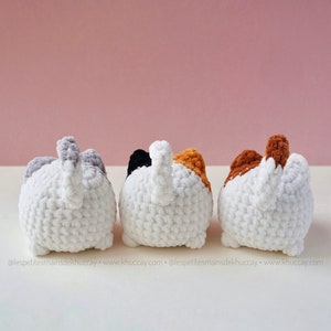 May include: Three crocheted cat toys, each with a different coloured tail. The cats are white with a grey, black, and orange tail, respectively. The cats are facing away from the camera.