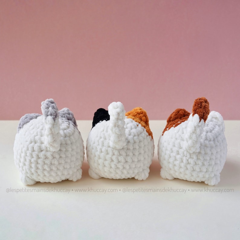 May include: Three crocheted cat toys, each with a different coloured tail. The cats are white with a grey, black, and orange tail, respectively. The cats are facing away from the camera.