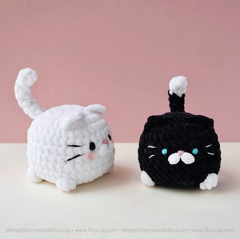 May include: Two crocheted cat plushies, one white and one black, with embroidered facial features. Both plushies have a white tail and are sitting on a white surface.