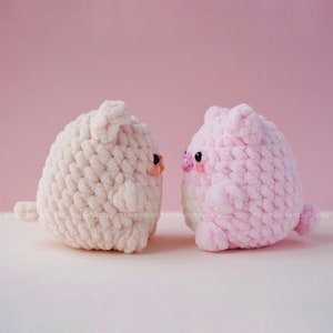 May include: Two crocheted pig plushies, one pink and one cream, facing each other against a pink background.