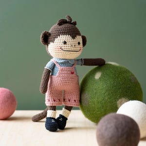 Peut inclure: Un singe en crochet marron portant une tenue bleue et rose se tient à côté d'une balle en feutre verte. Il y a aussi deux autres balles en feutre, une blanche et une marron.