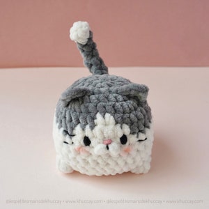 May include: A grey and white crocheted cat plush toy with a fluffy texture. The cat has a white face with black eyes and a pink nose.