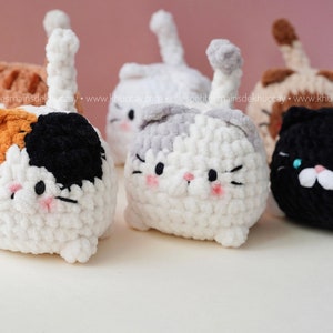 May include: Five crocheted cat plushies in different colours, including white, grey, black, orange, and brown. The plushies have black eyes, pink cheeks, and white noses. They are all sitting on a white surface.