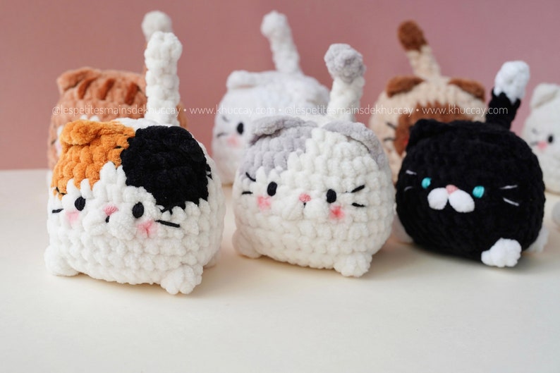 May include: Five crocheted cat plushies in different colours, including white, grey, black, orange, and brown. The plushies have black eyes, pink cheeks, and white noses. They are all sitting on a white surface.