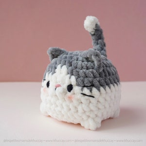 May include: A grey and white crocheted cat plush toy with a cute expression. The cat has a white face with black eyes and a pink nose. The cat's body is mostly grey with a white tummy.