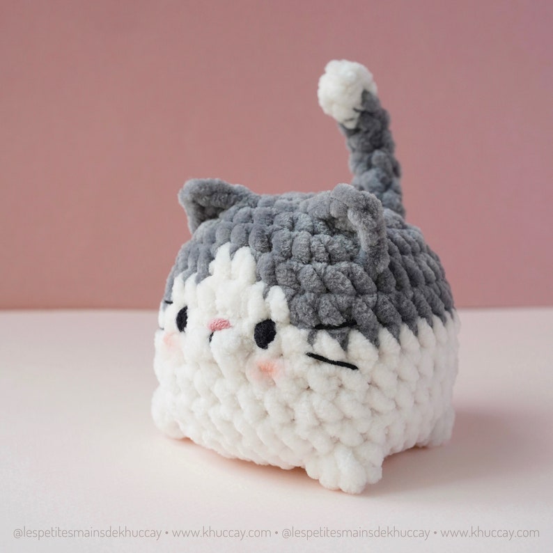 May include: A grey and white crocheted cat plush toy with a cute expression. The cat has a white face with black eyes and a pink nose. The cat's body is mostly grey with a white tummy.