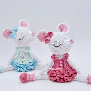 May include: Two crocheted mouse dolls wearing dresses and shoes. The mouse on the left is wearing a light blue dress with a light green bodice and a light blue bow. The mouse on the right is wearing a pink dress with a pink bow. Both mice have closed eyes and white fur.