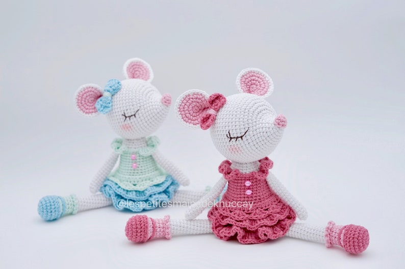 May include: Two crocheted mouse dolls wearing dresses and shoes. The mouse on the left is wearing a light blue dress with a light green bodice and a light blue bow. The mouse on the right is wearing a pink dress with a pink bow. Both mice have closed eyes and white fur.
