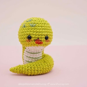 Puede incluir: Una serpiente verde de crochet con rayas blancas, ojos negros y boca roja. La serpiente tiene una apariencia linda y de dibujos animados.