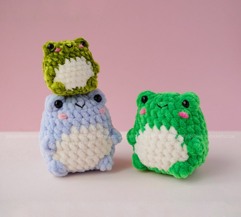 May include: Three crocheted frog plushies. The largest frog is green, the middle frog is blue, and the smallest frog is green. All three frogs have white bellies.