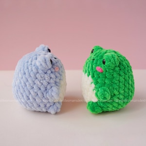 May include: Two crocheted plush toys shaped like frogs. One frog is light blue and the other is green. Both frogs have white bellies and pink cheeks.