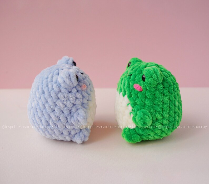 May include: Two crocheted plush toys shaped like frogs. One frog is light blue and the other is green. Both frogs have white bellies and pink cheeks.