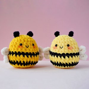 May include: Two crocheted bumblebees with black and yellow stripes and white wings. The bumblebees have smiling faces with black eyes and pink cheeks.