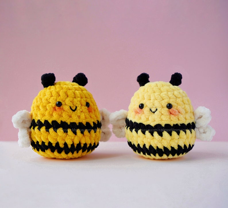 May include: Two crocheted bumblebees with black and yellow stripes and white wings. The bumblebees have smiling faces with black eyes and pink cheeks.