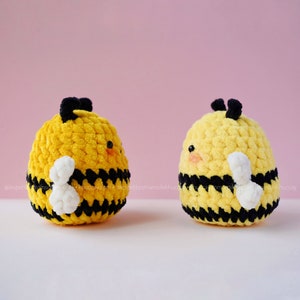 May include: Two crocheted bee plushies with black and yellow stripes and white wings. The bees have black eyes and a small, pink nose.
