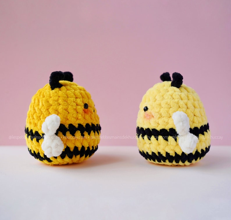 May include: Two crocheted bee plushies with black and yellow stripes and white wings. The bees have black eyes and a small, pink nose.