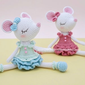May include: Two crocheted mouse dolls wearing dresses and ballet shoes. The mouse on the left is wearing a light blue dress with a pink bow, and the mouse on the right is wearing a pink dress with a pink bow. Both mice have closed eyes.