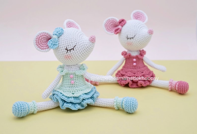 May include: Two crocheted mouse dolls wearing dresses and ballet shoes. The mouse on the left is wearing a light blue dress with a pink bow, and the mouse on the right is wearing a pink dress with a pink bow. Both mice have closed eyes.