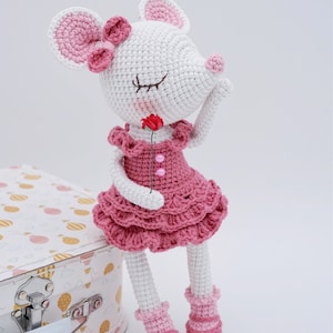 May include: A crocheted pink and white mouse doll wearing a pink dress and holding a pink flower. The doll is sitting on a white suitcase with a pink and yellow pattern.
