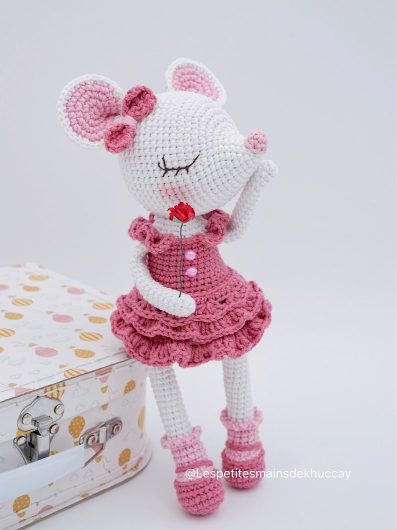 May include: A crocheted pink and white mouse doll wearing a pink dress and holding a pink flower. The doll is sitting on a white suitcase with a pink and yellow pattern.