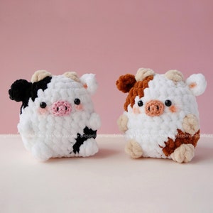 May include: Two crocheted stuffed cows, one black and white and one brown and white. Both cows have pink noses and black eyes.