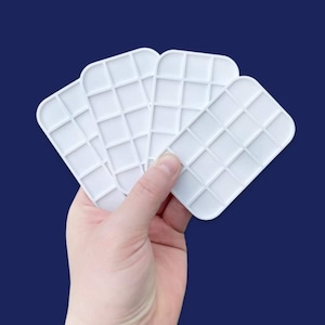 May include: Four white plastic trays with a grid pattern, held in a hand. The trays are designed for organising small items.