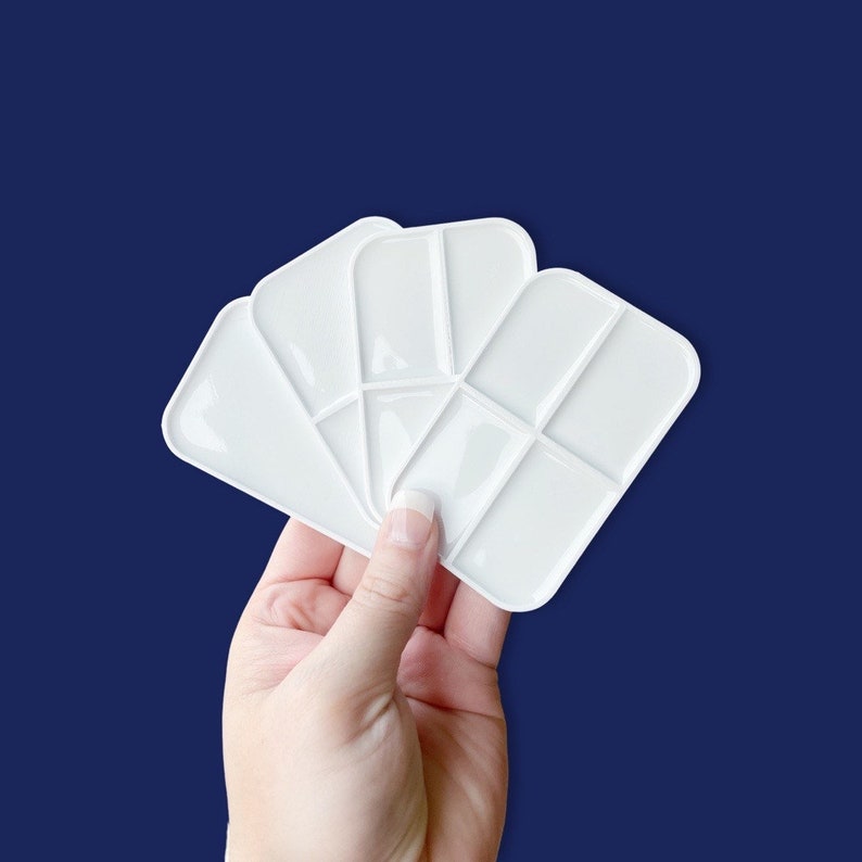 May include: A hand holding four white paint palettes. Each palette is rectangular with rounded edges and divided into sections for mixing colours. The background is a dark blue.