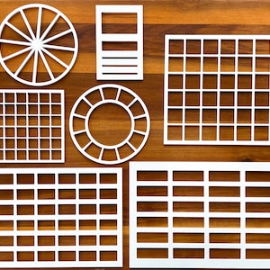 May include: A collection of white geometric stencils featuring various patterns including a circular wheel, a rectangular grid, a square grid, a circular frame, and two rectangular grids with horizontal slots, all arranged on a wooden surface.