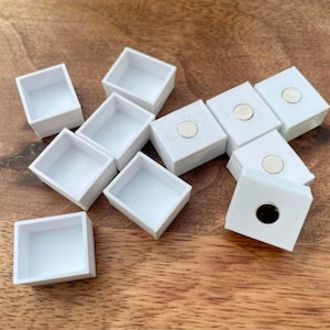 以下が含まれることがあります： 磁気閉鎖式の白い正方形のペイントパン10個セット。パンは木製の表面に緩い円形に配置されています。