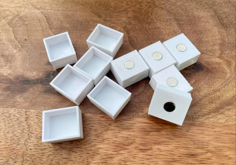 以下が含まれることがあります： 磁気閉鎖式の白い正方形のペイントパン10個セット。パンは木製の表面に緩い円形に配置されています。