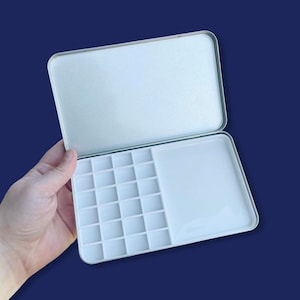 May include: A white metal paint palette with 18 individual paint wells and a larger well for mixing. The palette is open and has a white foam lining on the lid.