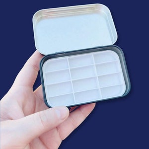 May include: A black metal tin with a white plastic insert containing 12 empty paint wells. The tin is open and the lid is off.