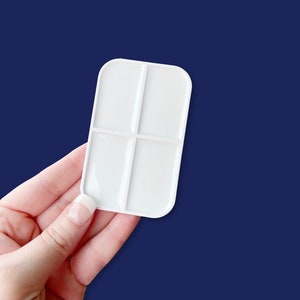 May include: A white rectangular paint palette with four sections, held against a dark blue background. The palette has rounded corners and is made of a smooth, glossy material. It is designed for mixing paints and is a useful tool for artists.