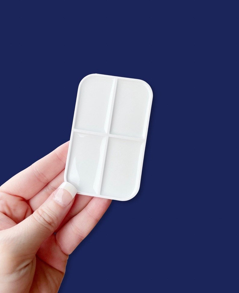 May include: A white rectangular paint palette with four sections, held against a dark blue background. The palette has rounded corners and is made of a smooth, glossy material. It is designed for mixing paints and is a useful tool for artists.
