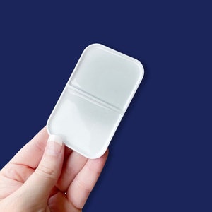 May include: A white, rectangular paint palette with rounded corners, divided into two sections. The palette is held against a dark blue background. The palette is designed for mixing paints and is a useful tool for artists.