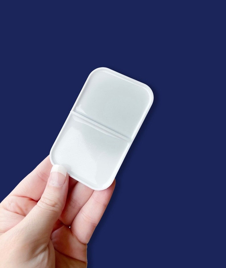 May include: A white, rectangular paint palette with rounded corners, divided into two sections. The palette is held against a dark blue background. The palette is designed for mixing paints and is a useful tool for artists.