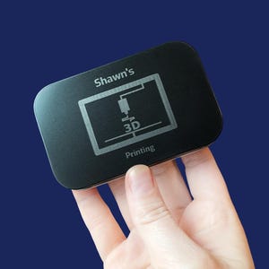 May include: A black rectangular object with rounded corners, held against a blue background. The object has the text "Shawn's" at the top and a 3D printing graphic with the word "Printing" below. The object appears to be a small, possibly electronic, device.