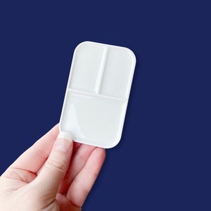 May include: A white paint palette with three compartments, held against a dark blue backdrop. The palette has a rectangular shape with rounded corners and is made of a smooth, glossy material.
