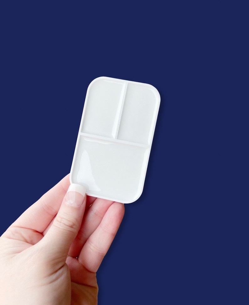 May include: A white paint palette with three compartments, held against a dark blue backdrop. The palette has a rectangular shape with rounded corners and is made of a smooth, glossy material.