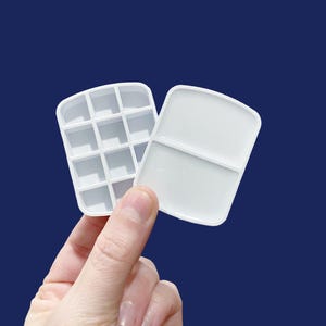 May include: Two white plastic paint palettes, one with 12 individual wells and the other with two larger wells.