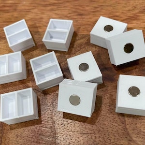 以下が含まれることがあります： 磁気閉鎖式の白いプラスチック製の容器が10個あります。一部の容器には2つの区画があり、他の容器には1つの区画があります。容器は木製の表面に配置されています。