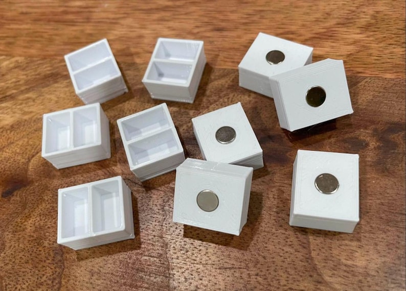 以下が含まれることがあります： 磁気閉鎖式の白いプラスチック製の容器が10個あります。一部の容器には2つの区画があり、他の容器には1つの区画があります。容器は木製の表面に配置されています。