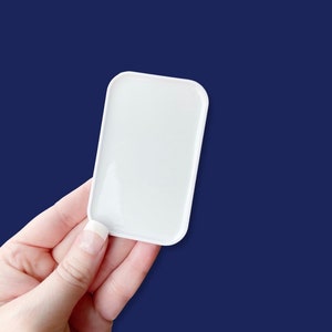 May include: A small, rectangular white tray with rounded corners is held against a dark blue background. The tray is smooth and glossy, reflecting light. The image focuses on the tray, highlighting its simple, clean design.