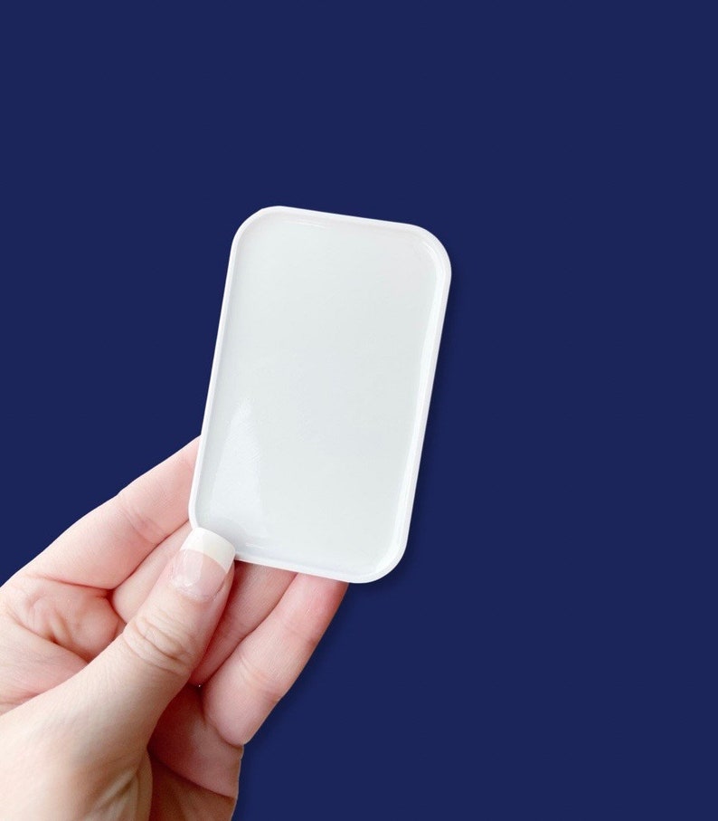 May include: A small, rectangular white tray with rounded corners is held against a dark blue background. The tray is smooth and glossy, reflecting light. The image focuses on the tray, highlighting its simple, clean design.