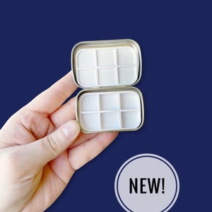 May include: A silver metal tin with a white plastic insert. The tin has 9 compartments and is open to reveal the empty compartments. The text "NEW!" is printed on a white circle in the lower right corner of the image.