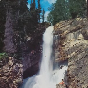 May include: A scenic postcard depicting a waterfall cascading down a rocky cliffside. Two people are standing on rocks near the base of the waterfall, enjoying the view.